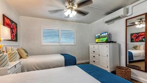 A bedroom with two beds, a ceiling fan, a TV, a dresser, and a window with blinds. - Menehune Shores #220 – Beachside Bliss with Panoramic Ocean Views (Kihei, Maui)