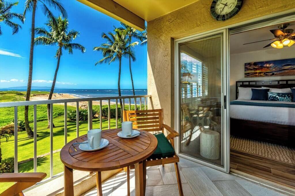 A balcony with a view of the beach and palm trees, a bed with a view, and a clock on the wall.
