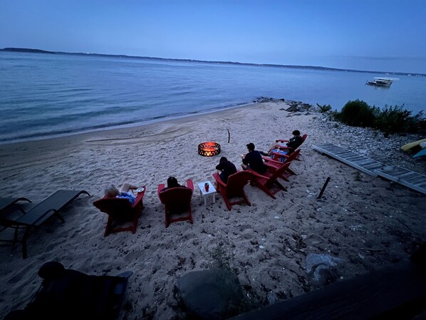 A beach with red chairs arranged in a row facing the water, a small fire pit in the center, and a distant boat on the water.
