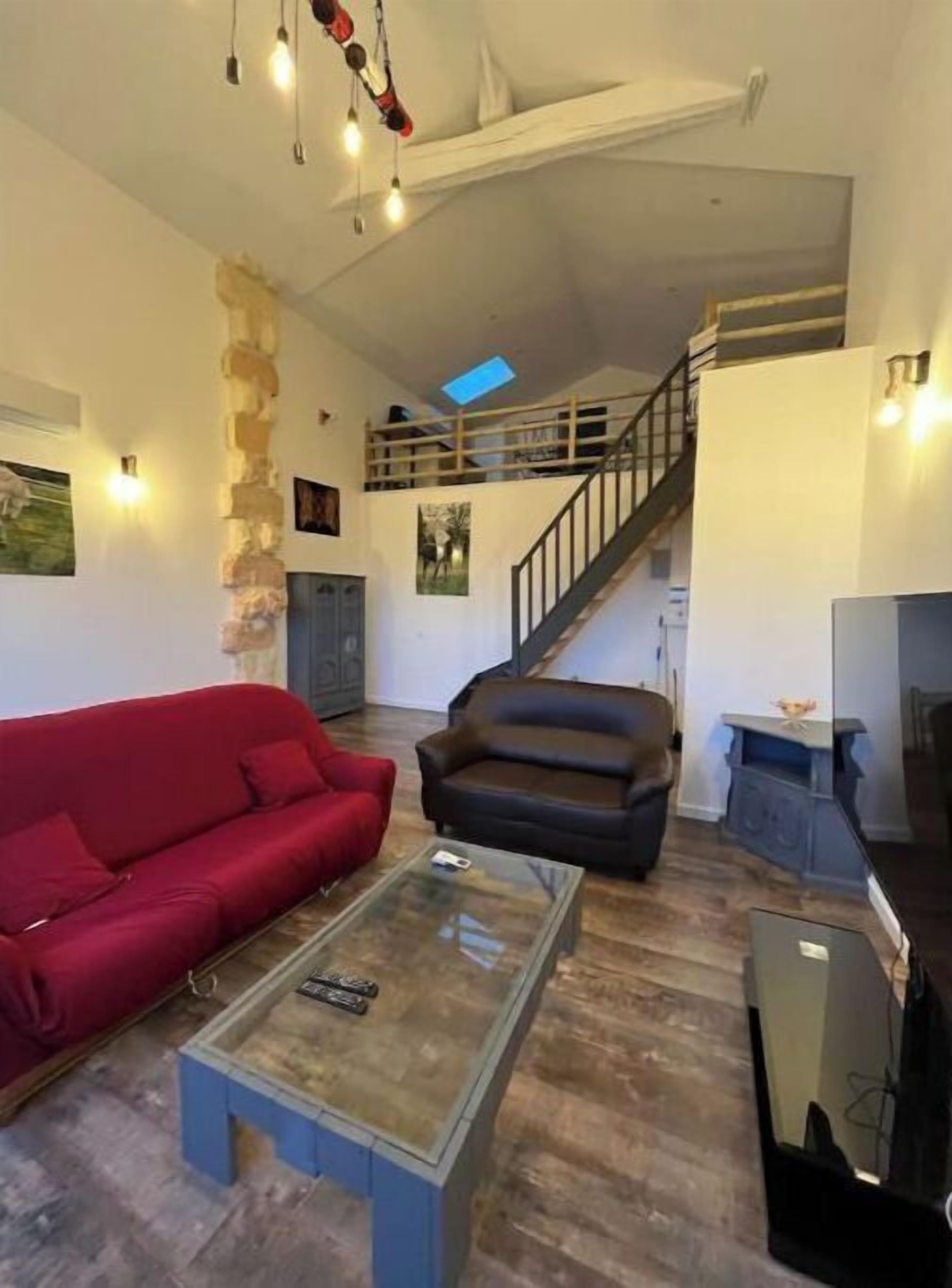 Ein geräumiger Wohnbereich mit einem roten Sofa, einem schwarzen Sessel und einem Glastisch. Eine Treppe führt auf eine obere Ebene, und ein Oberlicht erhellt den Raum.
