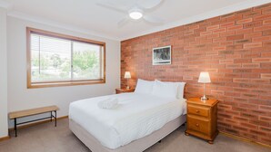 Ein Schlafzimmer mit einem Einzelbett, einer Bank, zwei Nachttischen mit Lampen, einem Deckenventilator, einem Fenster mit Jalousien und einer Ziegelwand.