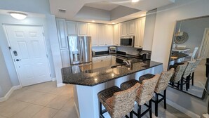 A modern kitchen with white cabinets, stainless steel appliances, a granite countertop, and a bar area with wicker chairs. - Escape to your poolside penthouse! (Bonita Springs)