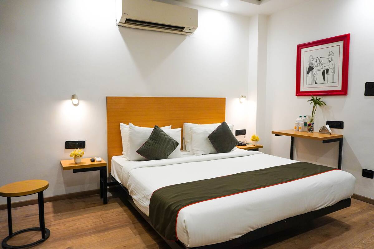 A hotel room with a bed, bedside tables, a wall-mounted air conditioner, a framed picture, and a small plant.