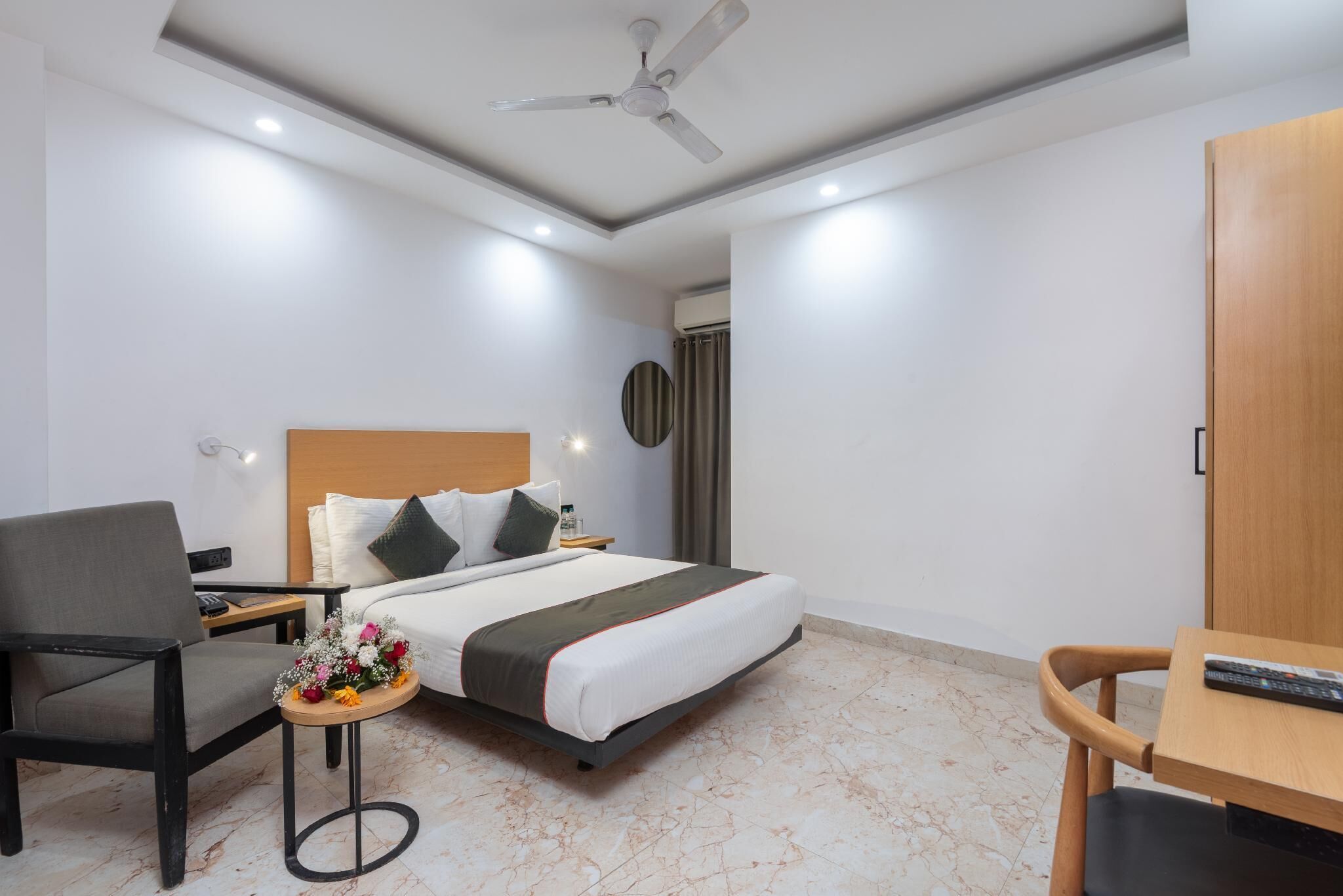 Une chambre d’hôtel comprenant un lit, un bureau, une chaise, une petite table avec un vase de fleurs, un ventilateur de plafond et un téléviseur fixé au mur.