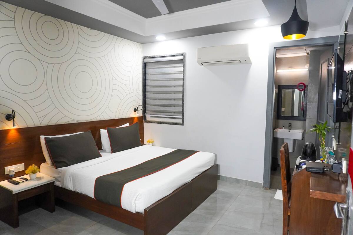 A hotel room with a bed, bedside tables, a nightstand, a desk, a mirror, an air conditioning unit, and a wall with a circular pattern.