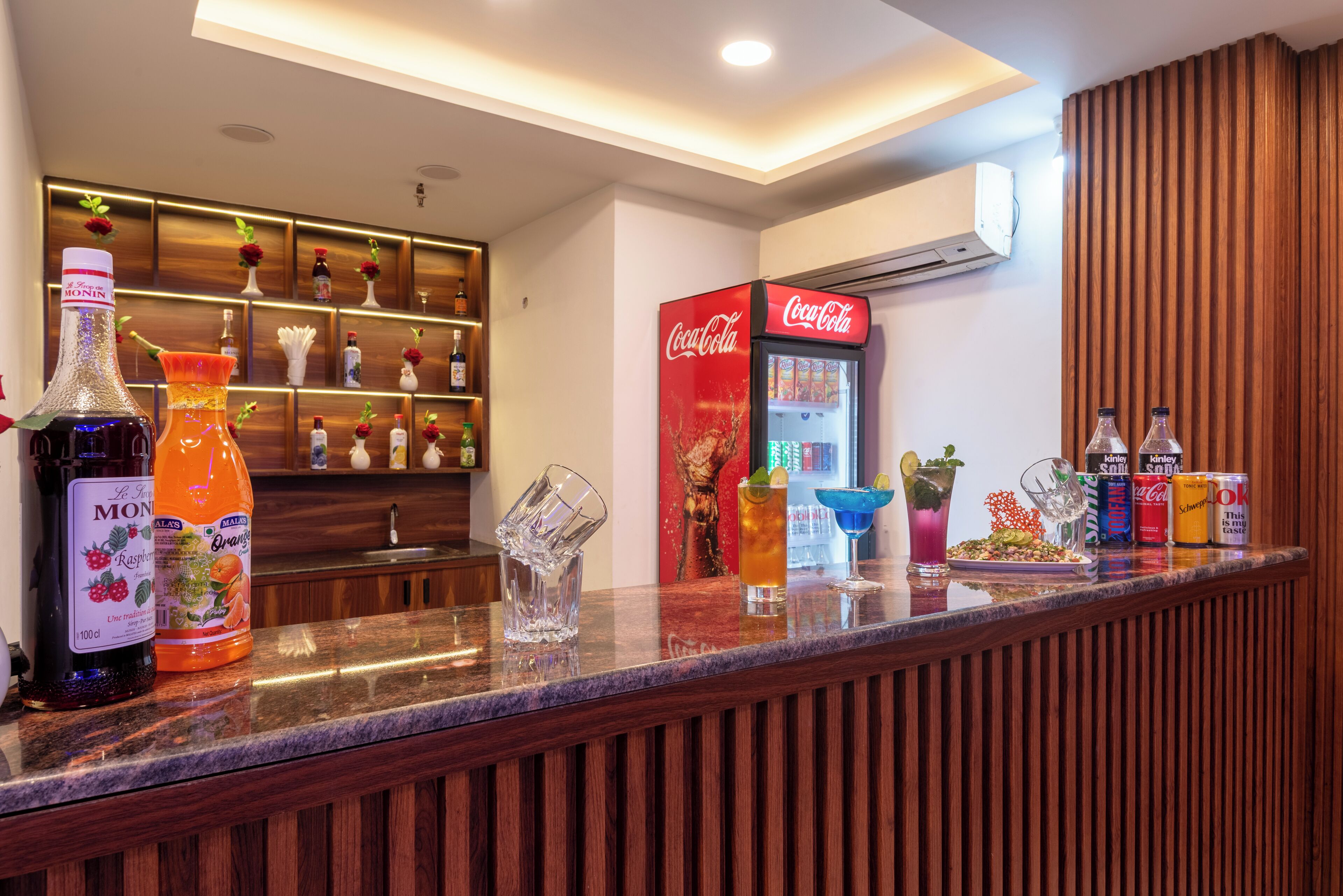 A bar area with a variety of beverages, including a Coca-Cola refrigerator and bottles of Monin syrup.