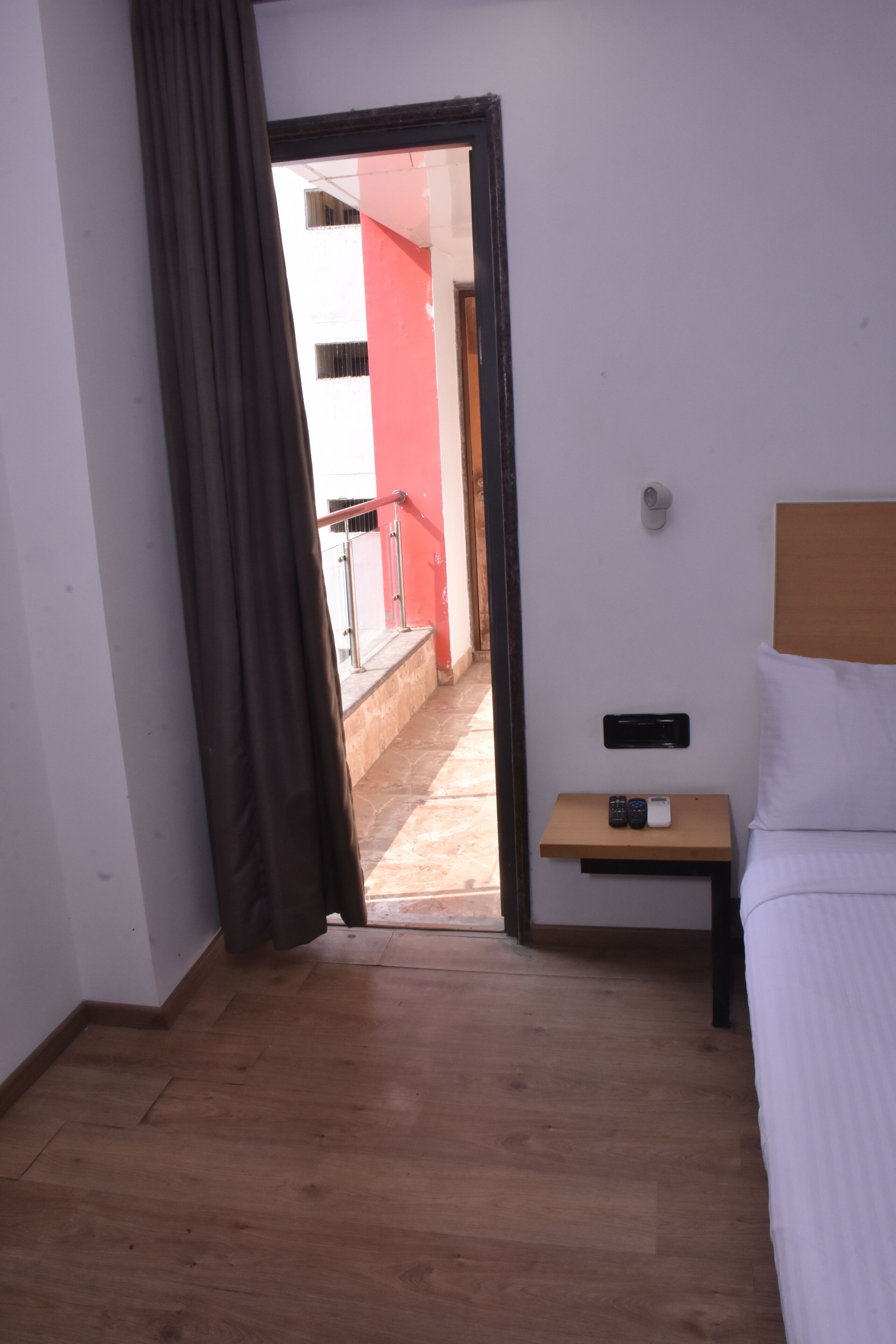 A hotel room with a wooden floor, a bed with white bedding, a nightstand with a remote control, and a view of a red building through an open door.