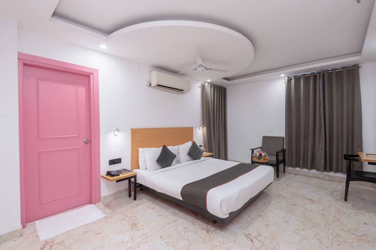 A hotel room with a pink door, a bed with white bedding and a dark throw, a desk with a chair, and a ceiling fan.