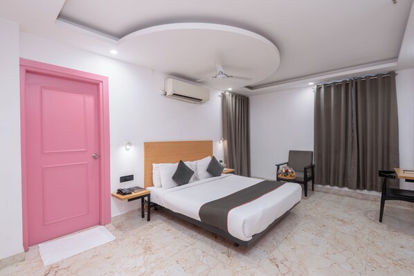 A hotel room with a pink door, a bed with white bedding and a dark throw, a desk with a chair, and a ceiling fan. - Chhavi Hotels (Noida)