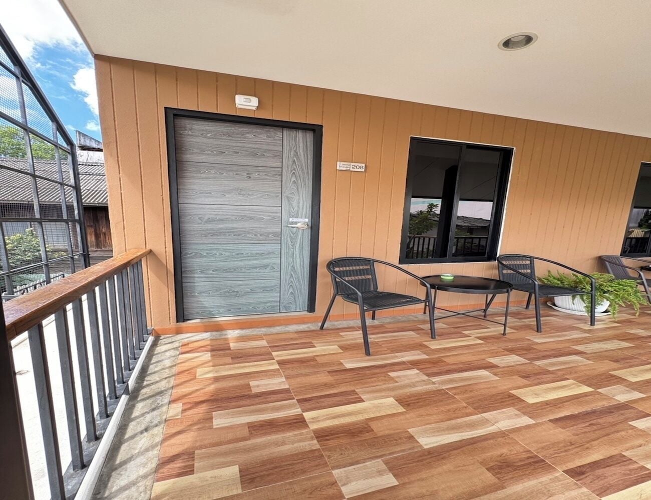 A balcony with a wooden floor, a metal railing, and a door. There are two chairs and a table with a plant on the balcony.