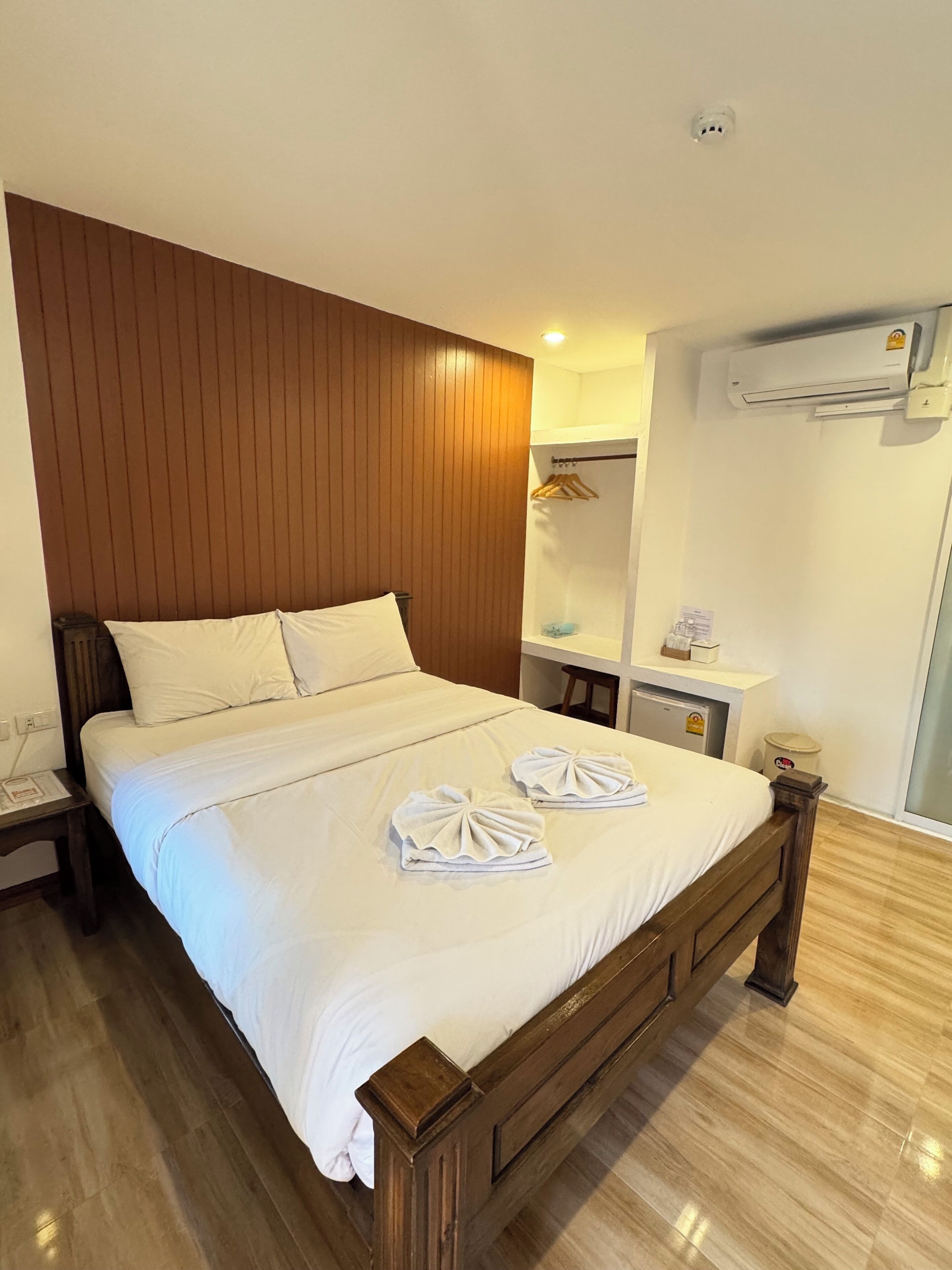 A hotel room with a wooden headboard, a large bed with white linens, two towels on the bed, a bedside table, a small desk with a chair, an air conditioning unit, and a mirror on the wall.