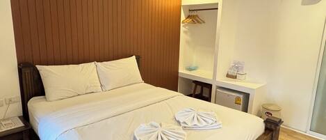 A hotel room with a wooden headboard, a large bed with white linens, two towels on the bed, a bedside table, a small desk with a chair, an air conditioning unit, and a mirror on the wall.