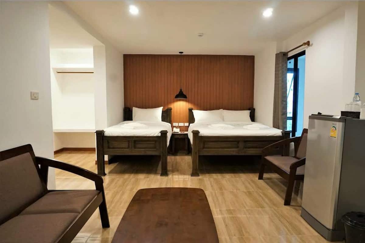 Quarto de hotel com duas camas, um sofá, uma mesinha, uma geladeira e uma parede de destaque em madeira.