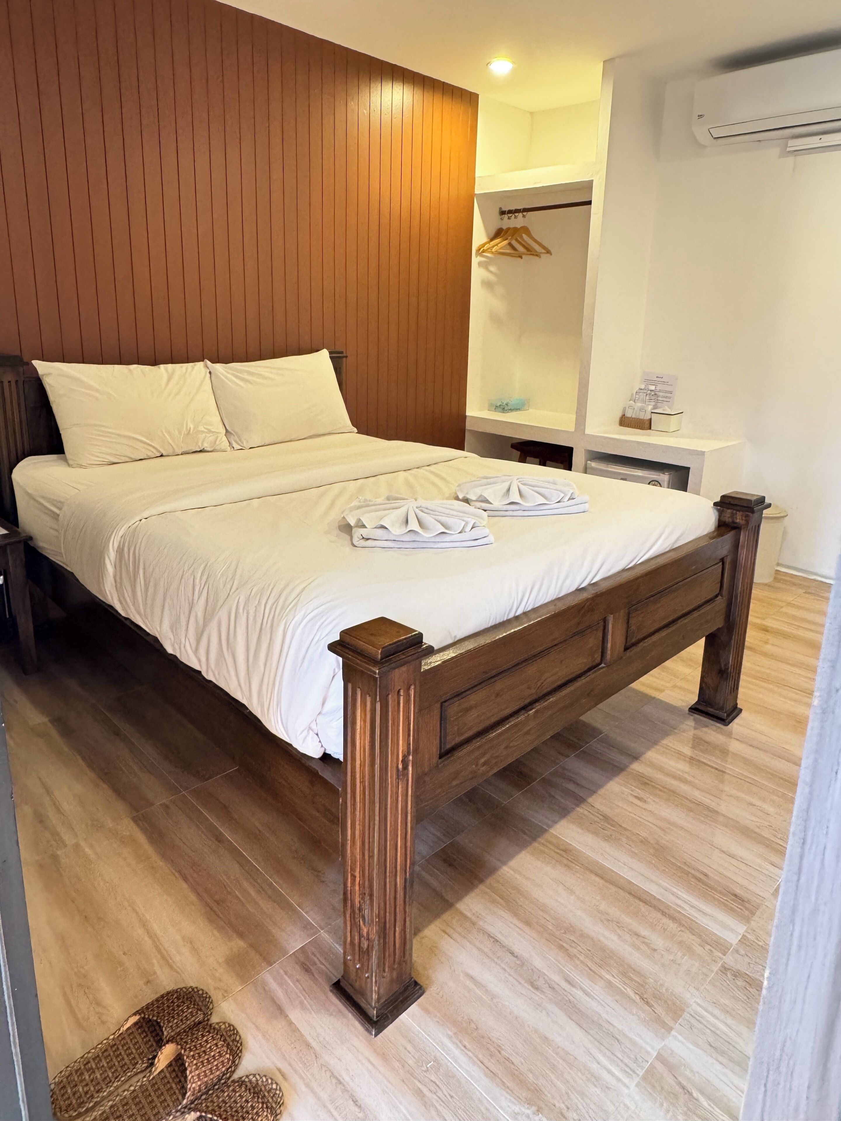 Ein ordentlich bezogenes Bett mit Holz Kopfteil, ein kleiner Nachttisch mit Lampe und eine Wand-Klimaanlage.