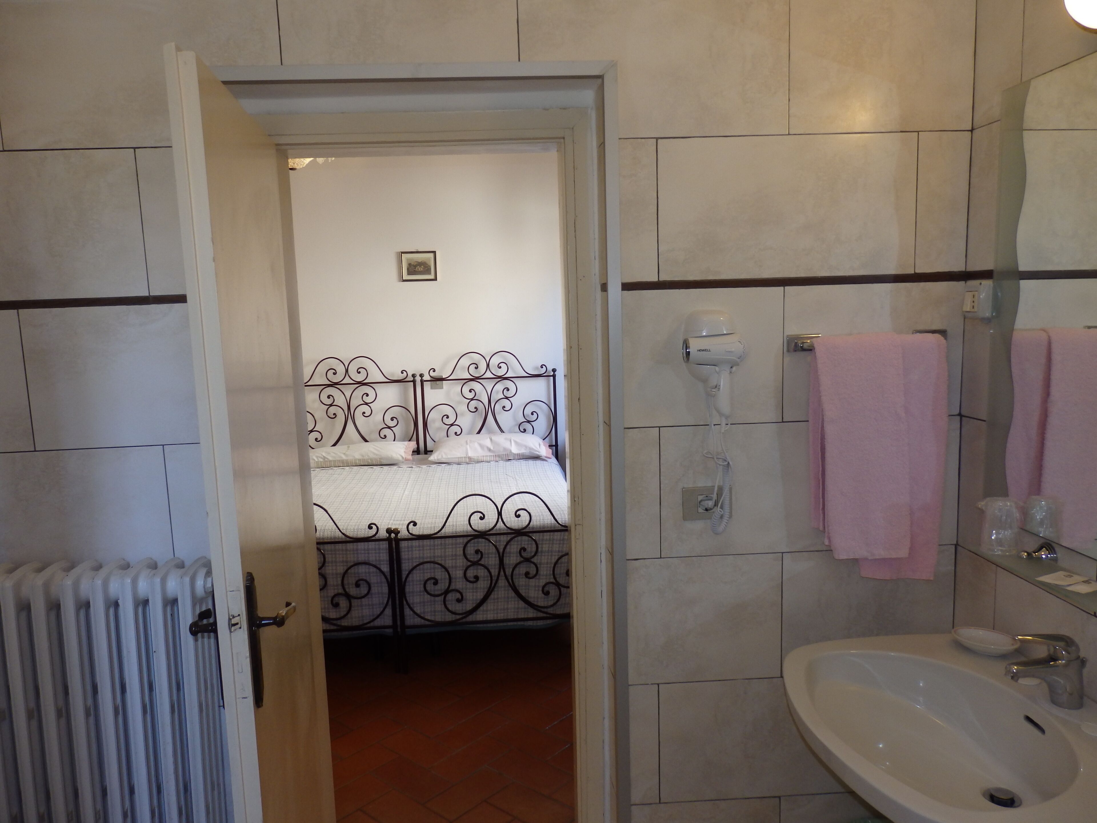 Une chambre à coucher avec un lit en métal, un radiateur blanc et une salle de bain équipée d’un lavabo et d’un porte-serviettes.