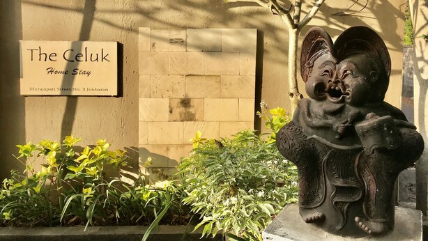 Segnale per 'The Celuk Home Stay' su una parete color beige, con una statua decorativa di un angelo in primo piano.