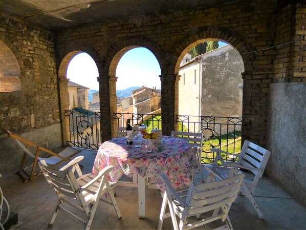 A round table with a floral tablecloth, surrounded by white chairs, set against a stone wall with arched openings.