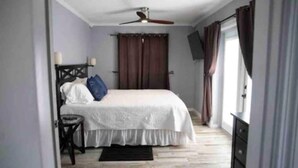 Een slaapkamer met een bed, een stoel, een televisie, een plafondventilator en een raam met gordijnen.