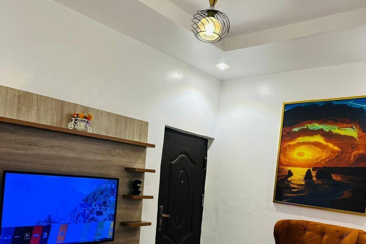 Una sala de estar con televisor de pantalla plana, un sofá, una silla, una mesita redonda con un florero y un cuadro en la pared.