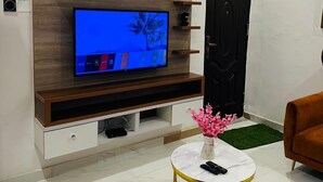 Uma sala de estar moderna com uma televisão de tela plana presa em um painel de madeira, uma mesa de centro redonda com um vaso de flores, e uma porta preta.