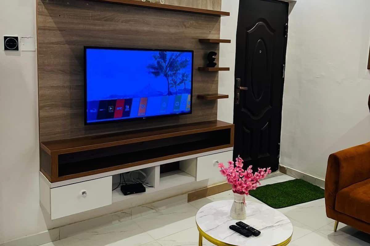 Una sala de estar moderna con un televisor de pantalla plana montado en un mueble de pared de madera, una mesa redonda con un jarrón de flores y una puerta negra.