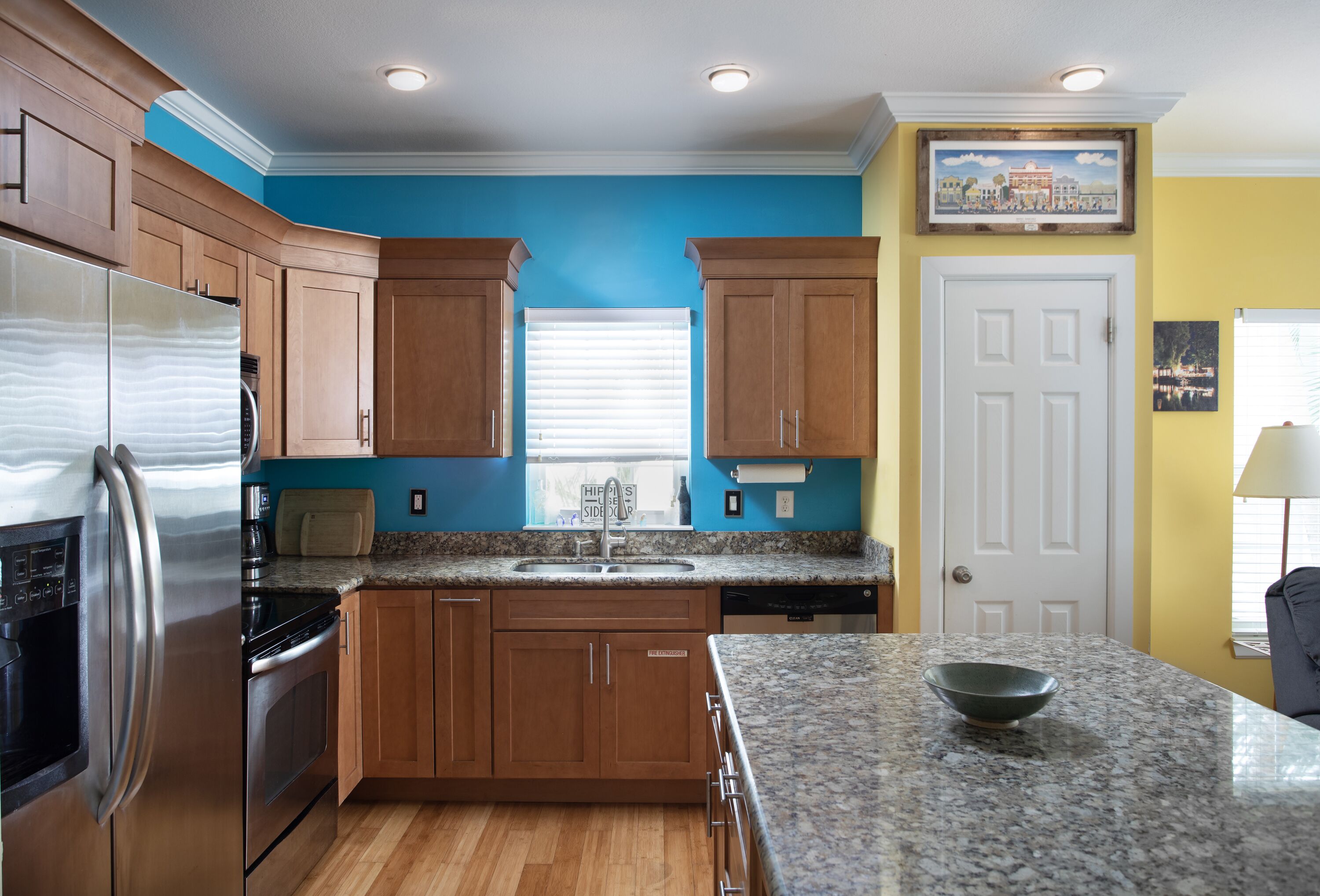 A kitchen with wooden cabinets, a stainless steel refrigerator, granite countertops, and a window with blinds.