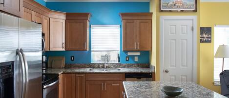 A kitchen with wooden cabinets, a stainless steel refrigerator, granite countertops, and a window with blinds.