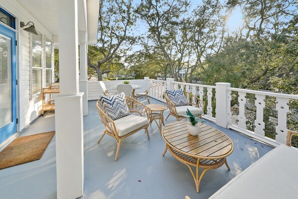 A covered outdoor seating area with wicker furniture and a round table, surrounded by a white railing and trees.