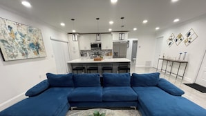 A modern living room with a blue sectional sofa, a glass coffee table with a plant, and a kitchen in the background. - Pool House 3/2: Grill, Mural and Ping Pong Table (Sunrise)