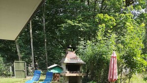 Een terras buiten met een stenen vloer, een houten picknicktafel, twee blauwe lounge stoelen, een barbecue en een roze parasol.
