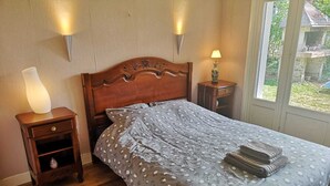 Een slaapkamer met een houten bed, een nachtkastje met een lamp en een raam met uitzicht op groen.
