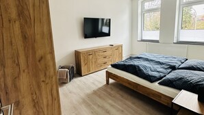 A bedroom with a wooden wardrobe, a bed with blue bedding, a TV mounted on the wall, and large windows. - Vacation apartment in Obernkirchen (Obernkirchen)