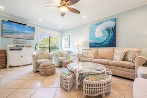 A living room with a large TV, a ceiling fan, a sofa, wicker furniture, and a beach-themed painting. - Golf Cart ~ Multiple Pools ~ Screened Back Patio (Miramar Beach)