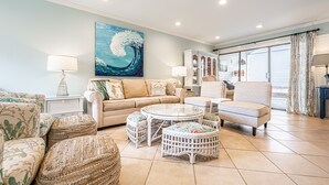A spacious living room with a large sofa, two armchairs, a coffee table, and wicker ottomans. There is a painting of a wave on the wall, a ceiling fan, and a large window with curtains. - Golf Cart ~ Multiple Pools ~ Screened Back Patio (Miramar Beach)
