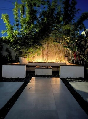 A modern outdoor seating area with a fire feature, surrounded by lush greenery and a pathway. - OASIS VILLAGE (Vega Baja)