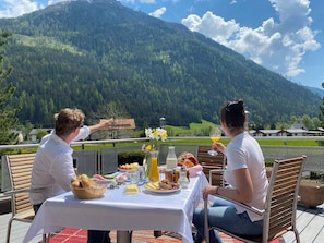 A table set outdoors with a mountain view, including a basket of bread, a glass of orange juice, and various condiments.