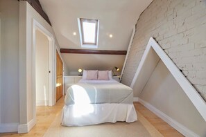 Quarto com teto inclinado, claraboia, cama com almofadas, abajures e piso de madeira.