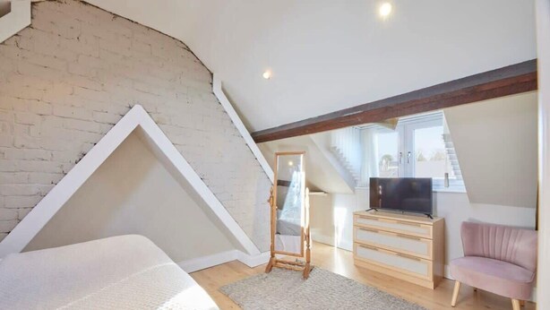 Een slaapkamer met een zolderkamer, een houten dressoir, een televisie, een roze stoel en een spiegel.