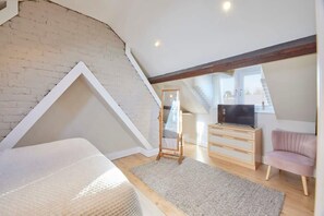 Une chambre à coucher aménagée dans un grenier, avec un plafond en pente, une commode en bois, un téléviseur, un fauteuil rose et un miroir.