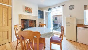 Een eetruimte met een ronde tafel en houten stoelen, een open haard, een keuken met een koelkast en een klok aan de muur.