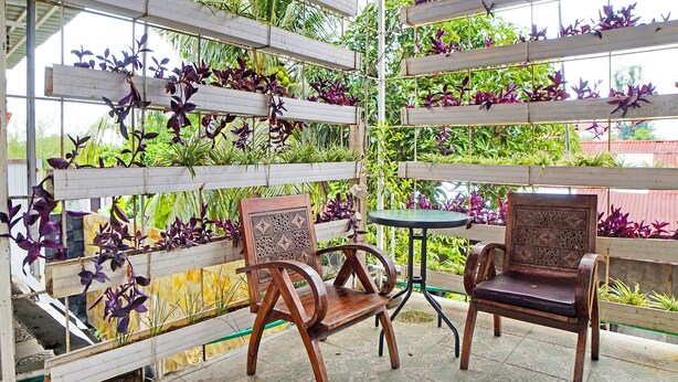 A covered outdoor seating area with wooden chairs and a small table, surrounded by vertical garden shelves with various plants.