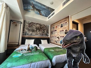 Une chambre d’hôtel avec deux lits, de la literie sur le thème des dinosaures, une fresque murale et une grande figurine de dinosaure.
