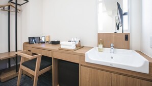 A hotel room with a wooden desk, a chair, a white sink, a mirror, and a towel rack.