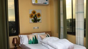 Een hotelkamer met een bed, nachtkastje, lamp, schilderij van zonnebloemen en een raam met gordijnen.