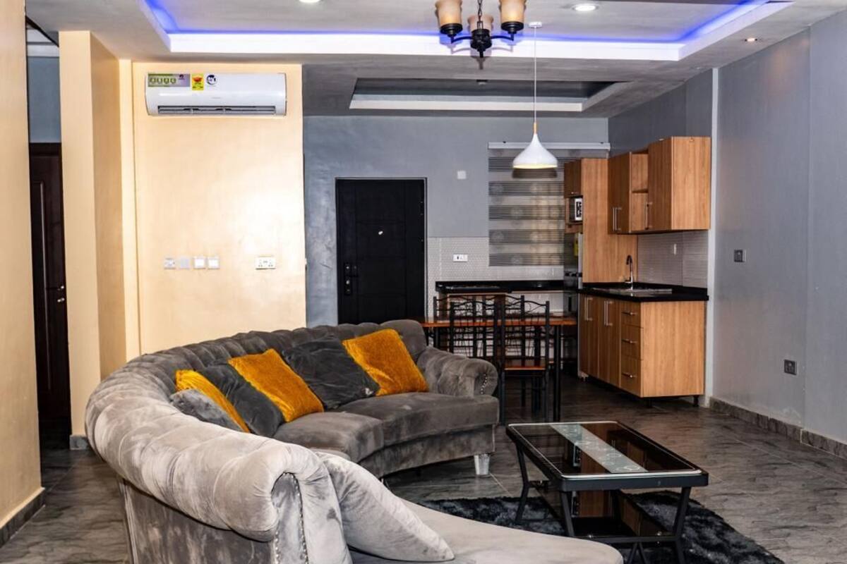 A modern living room with a grey sofa, a coffee table, and a kitchen area with wooden cabinets and a black countertop.