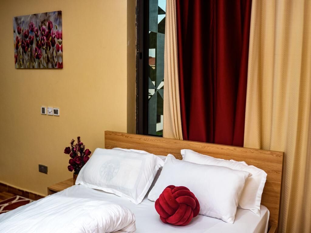 Habitación de hotel con cabecera de madera, ropa de cama blanca, un cojín decorativo rojo, un cuadro de flores en la pared y una ventana con cortinas rojas.