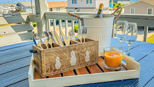 A wooden table with a tray containing a pineapple-themed knife block, a candle, and a glass of orange juice. In the background, there is a house with a balcony. - La Carolina del Mar - KDH MP 5.5, 250 Yds to Beach, Ocean View, Hot Tub, Flex Stays (Kill Devil Hills)