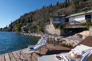 A wooden deck with lounge chairs and a table, overlooking a body of water and a hillside with buildings.