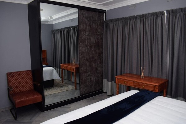 A hotel room with a large mirror, a bed with a dark blue bedspread, a wooden nightstand, a red upholstered chair, and grey curtains.