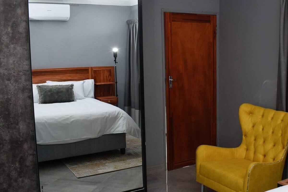 A bedroom with a large mirror, a bed, a wooden headboard, a nightstand, a yellow armchair, a wooden door, and a chandelier.